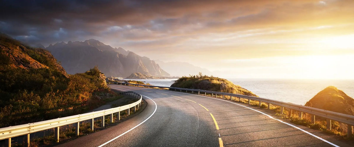 Picturesque road lined with greenery, leading to mountains and ocean.