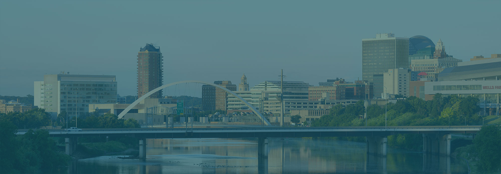 Modern city skyline with an arched bridge over a calm river surrounded by greenery under a clear sky.